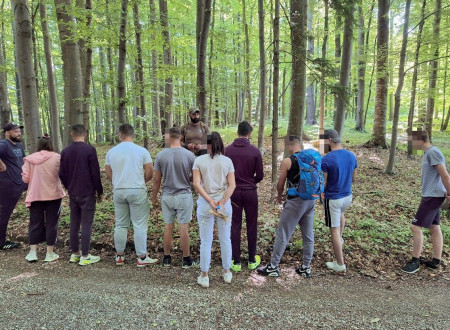 Skupina mladoletnikov, obrnjenih s hrbtom proti objektivu, posluša predstavitev v gozdu.
