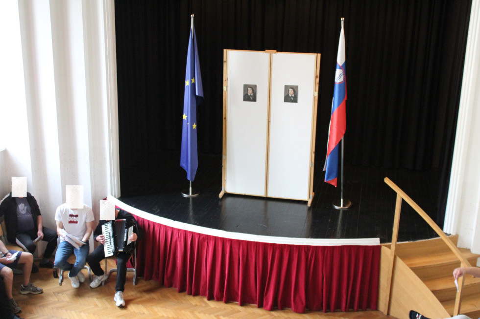 Oder in postavitev ob kulturnem prazniku. Dve sliki Franceta Prešerna, slovenska in evropska zastava. Pred odrom sedijo moški. Eden ima harmoniko v roki.