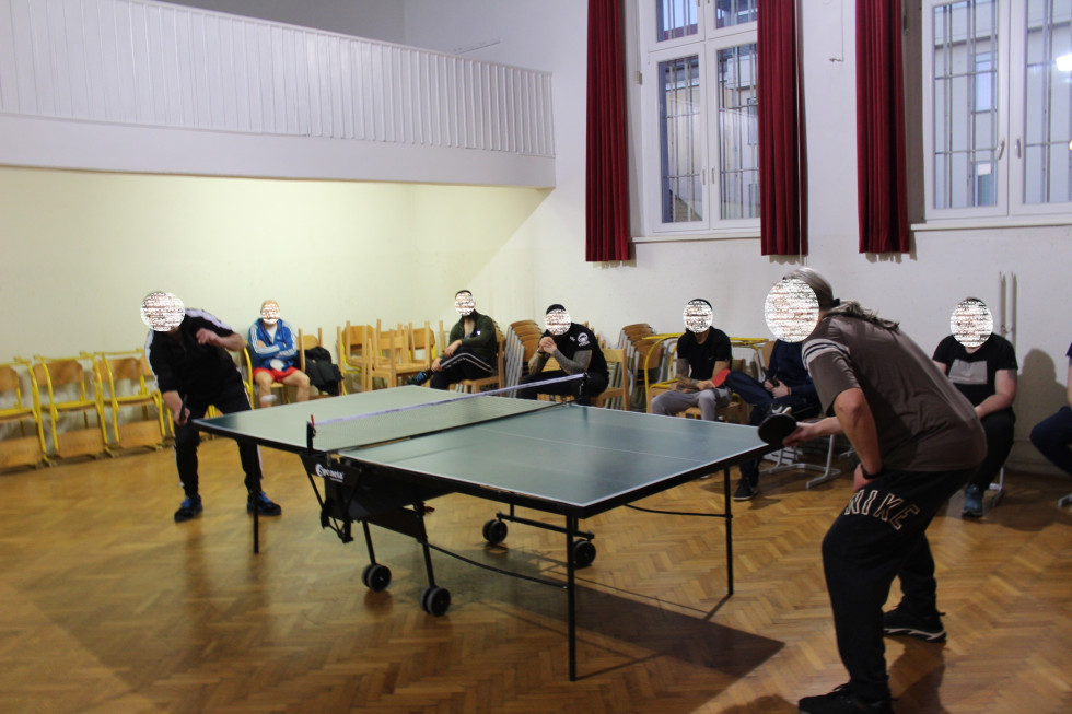 Udeleženci turnirja v namiznem tenisu, dva igralca vsak na svoji strani mize in ostali navzoči, ki opazujejo.