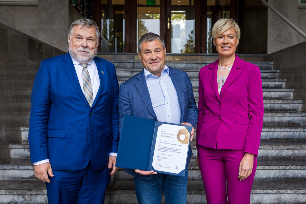 vsi trije stojijo pod vhodnimi stopnicami inštituta, nagrajenec je v sredini in kaže razprto nagrado v mapi