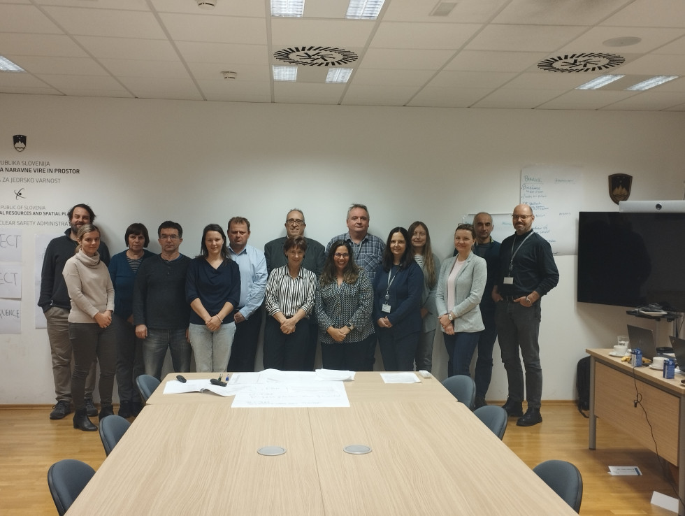 15 udeležencev stoje pred steno predavalnice, v ospredju stoli in mize