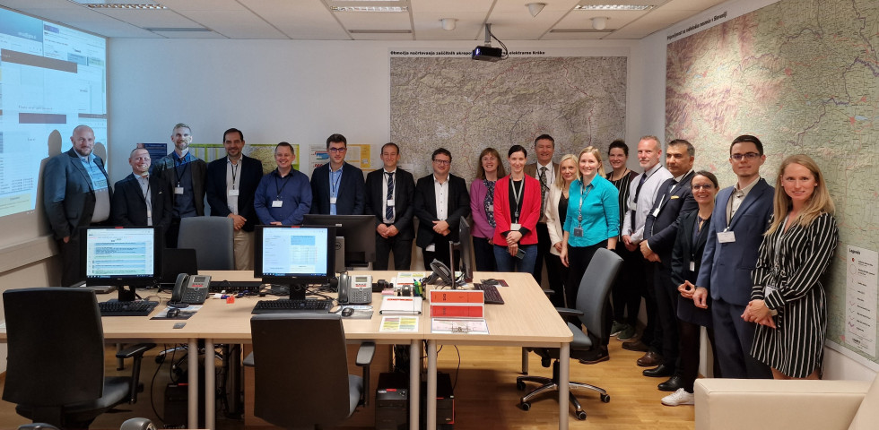 19 participants (12 men and 7 women) standing by the room wall. The walls are covered by maps and by projected computer screen. In front of the room are two office desks with two computers, two chairs and stationery equipment on the desks.