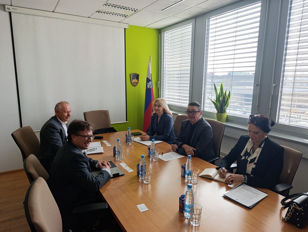 Trije moški in dve ženski sedijo za mizo. Na mizi papirji in voda. V kotu sobe slovenska zastava.