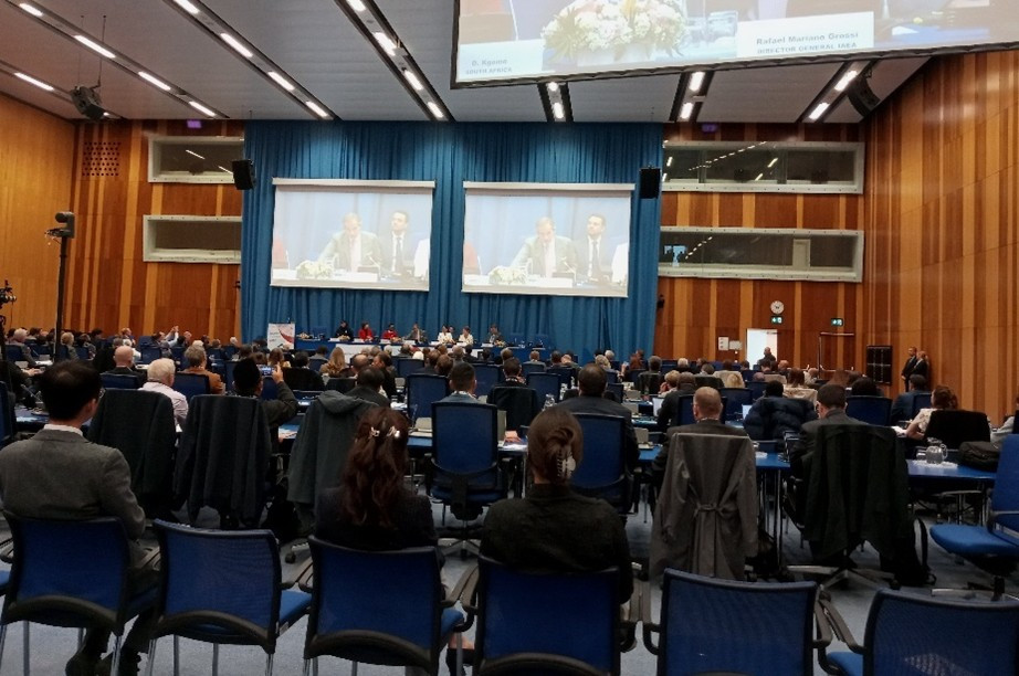 Konferenčna dvorana z udeleženci konference, ki poslušajo uvodni nagovor generalnega direktorja MAAE. Njegov nastop je viden tudi na dveh panojih. viden 