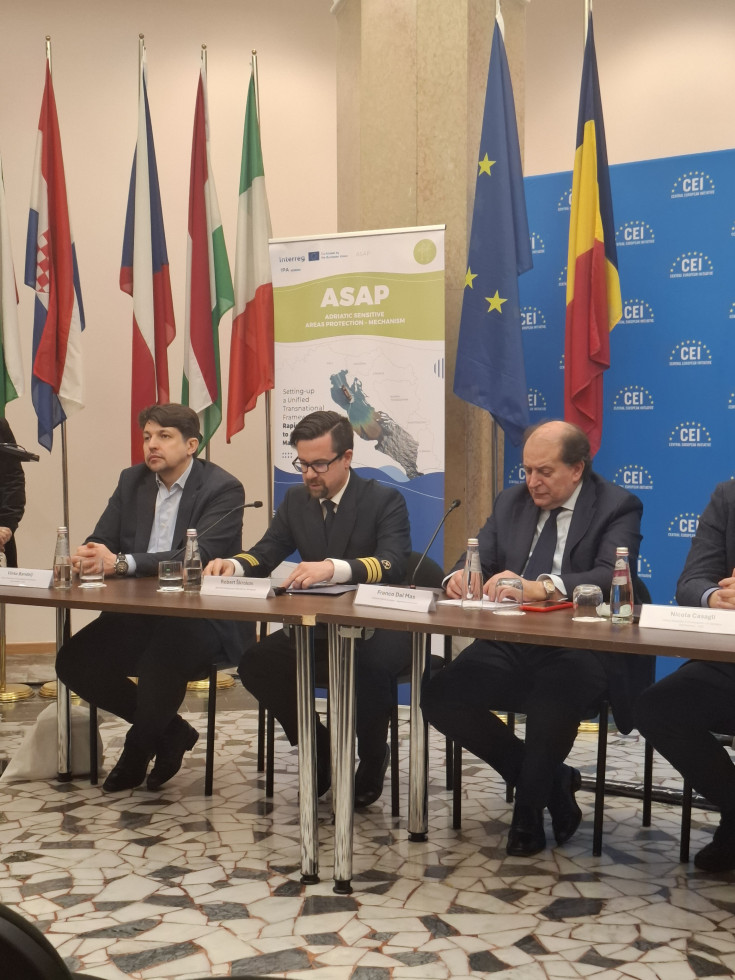 Govornik Robert Škrokov sedi za mizo. Zraven njega dva moška. Zadaj zastave.