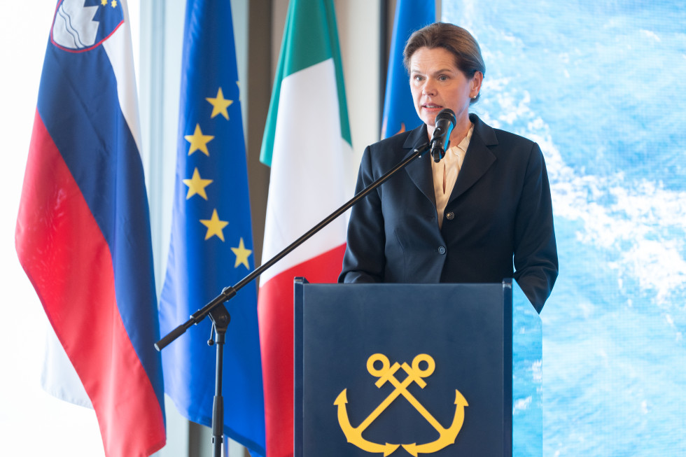 Ministrica za infrastrukturo, mag. Alenka Bratušek stoji za govorniškim pultom. Zadaj slovenska evropska in italijanska zastava.