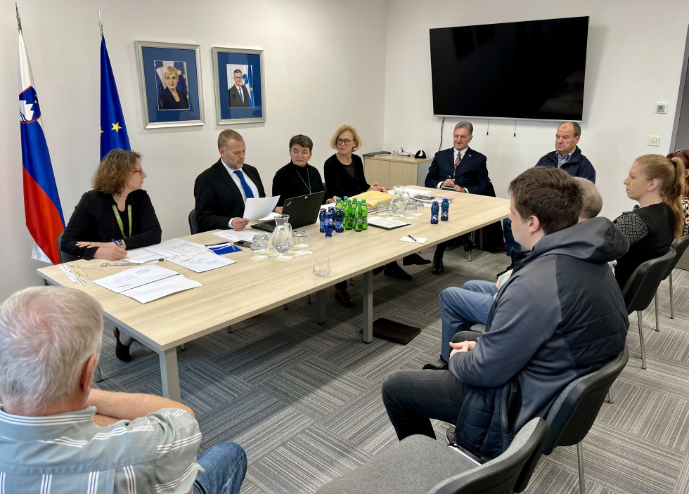 Člani komisije za odpiranje vlog sedijo za mizo pred udeleženci, v ozadju na steni sliki predsednice Republike Slovenije in ministra za obrambo