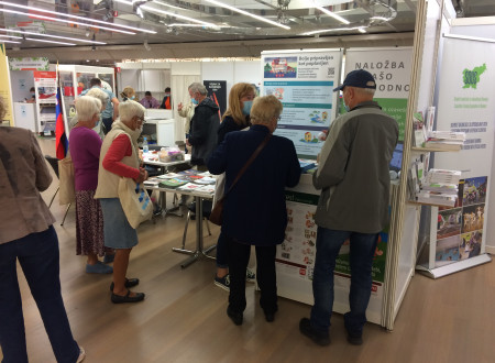 Obiskovalci si ogledujejo razstavni prostor Uprave Republike Slovenije za zaščito in reševanje. Na stojnici so med drugim tudi zgibanke na temo poplave in promocijski material. Viden je plakat na temo poplave in kohezijskega projekta.