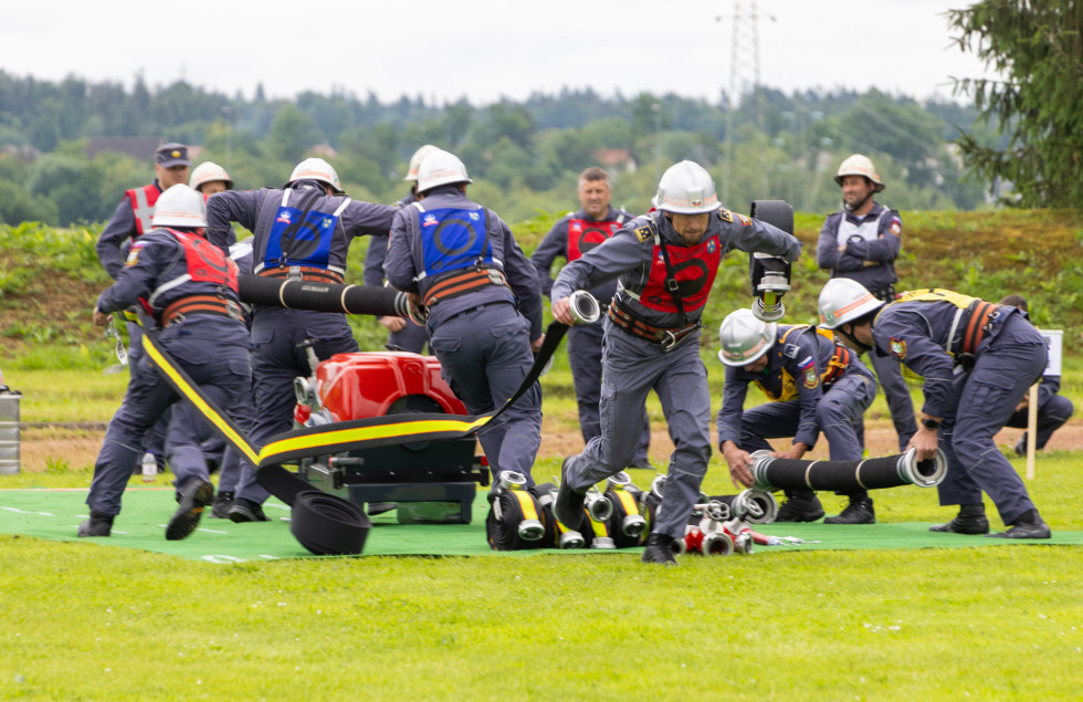 Slika prikazuje gasilce, ki tekmujejo v vlečenju brizgalne. 