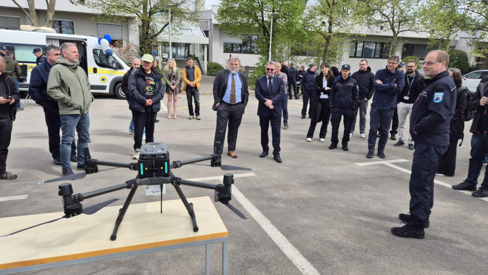 Na fotografiji je prikazan dron s posebnim detektorjem. Pred njim je zbrana skupina ljudi, ki si ogleduje ta dron. Med ljudmi sta dr. Leon Cizelj z Instituta Jožef Stefan in generalni direktor Uprave za zaščito in reševanje Leon Behin.