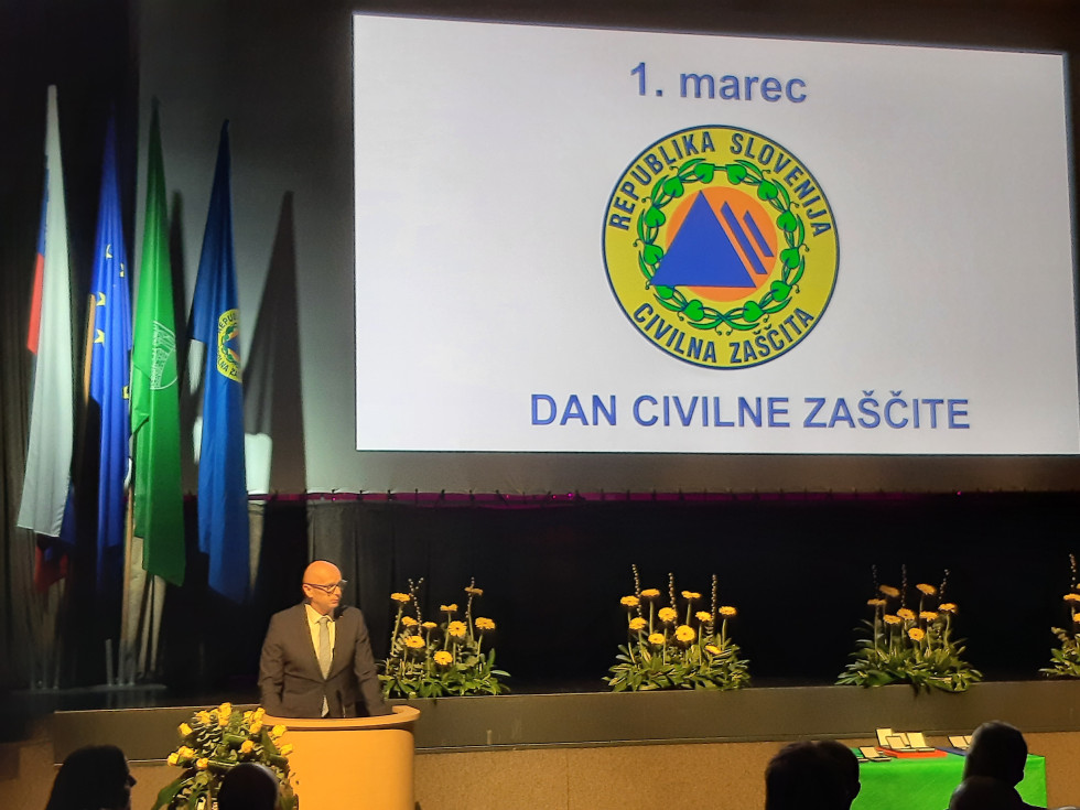 Moški stoji za govorniškim pultom. Za njim zastave in veliko platno s predstavitvijo. Napis: 1. marec; dan civilne zaščite.