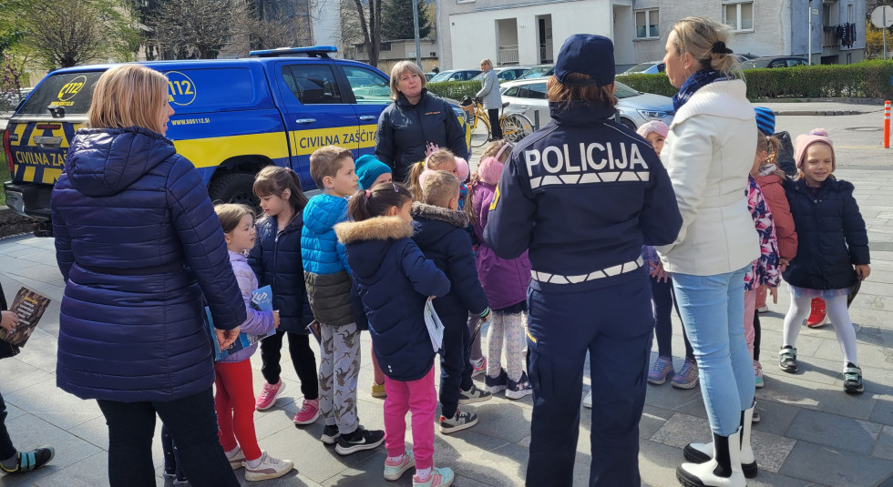 Policajka obrnjena s hrbtom proti nam, skupina otrok z vzgojiteljico ter predstavnica civilne zaščite v ozadju vozilo civilne zaščite in bloki.