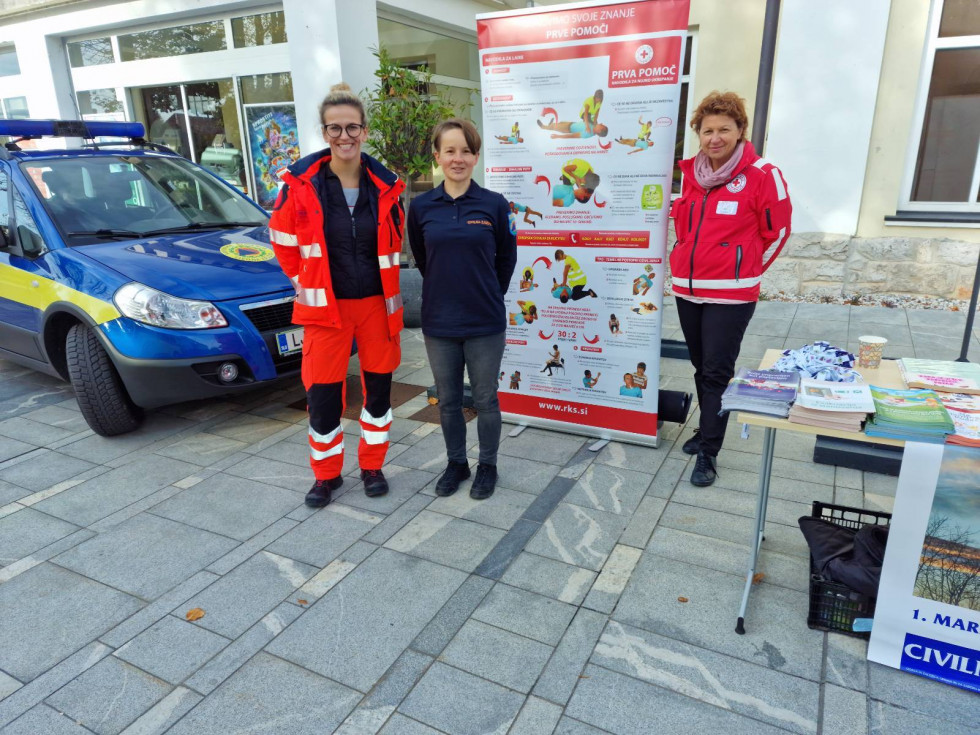 Predstavnica nujne medicinske pomoči, civilne zaščite in rdečega križa pred razstavnim panojem v ozadju vozilo modre barve.