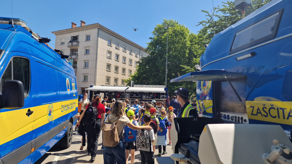 Na fotograiji je skupina otrok med dvema vozila civilne zaščite, v ozadju stolpnica. Otrok spremlja učiteljica, zbrani so v skupini.