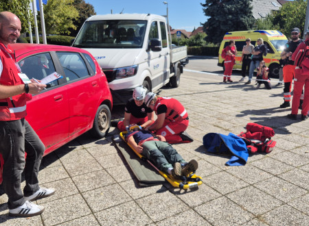 Na fotografiji je scenarij prometne nesreče, ocenjevalec stoji pri avtu, na tleh letži človek, ki so ga dali v nosila, ob njem je bolničar, v ozadju še reševalno vozilo