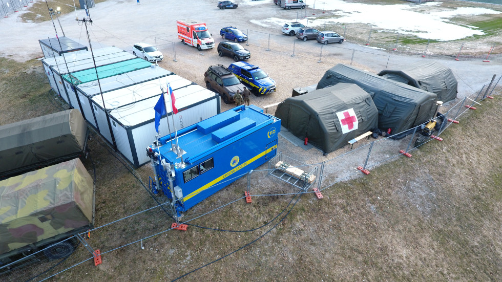 Kontejner modre barve z napisom Civilna zaščita, na njem zastavi EU in SLO, zraven kontejnerja še šotor Slovenske vojske in drugi kontejnerji. V zgornji tretjini slike vozila.