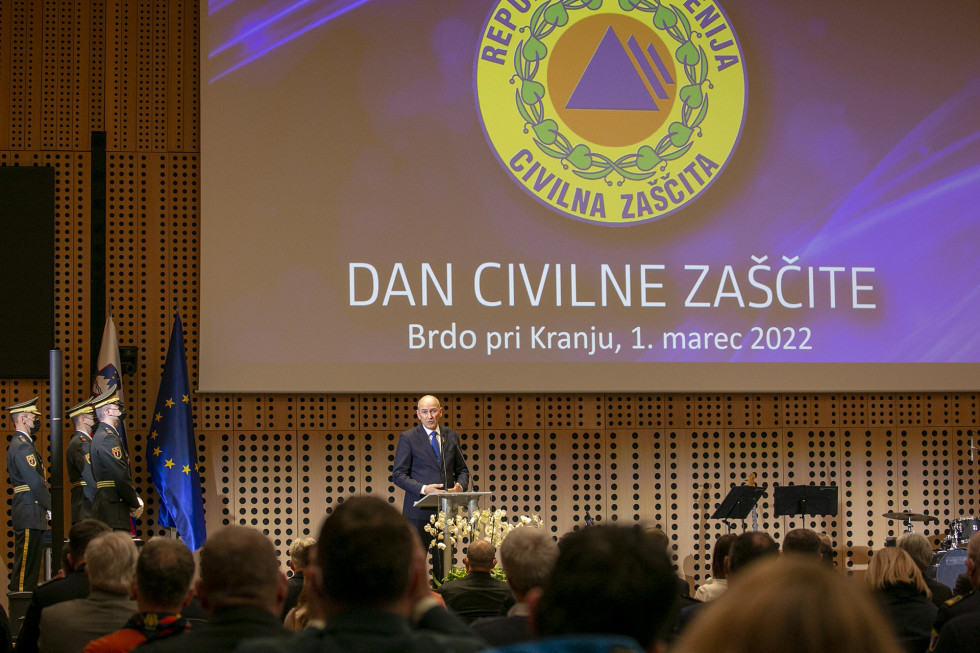 Za govorniškim pultom na odru predsednik Vlade, v ozadju platno z znakom Civilne zaščite in napisom dan Civilne zaščite. V ospredju udeleženci dogodka gledajo proti odru.