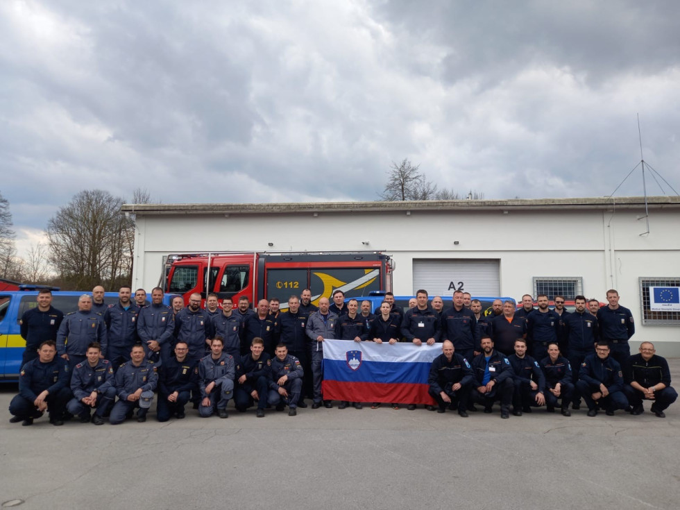 Prva vrsta čepi, druga vrsta stoji, v sredini slovenska zastava, v ozadju gasilsko vozilo in stavba.