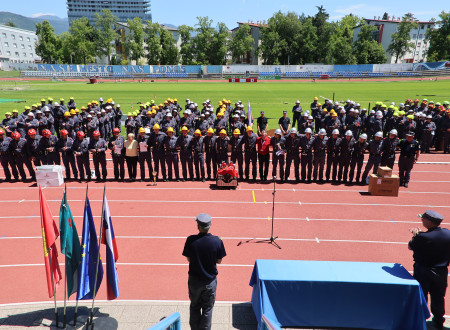Na fotografiji so postrojeni gasilci na stadionu, pred njimi so zastave  in govorniški mikrofon