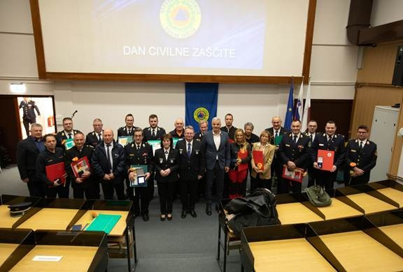 Na sredini slike so prejemniki priznanj, v zgornjem delu platno in na njem znak Civilne zaščite, v spodnjem delu klopi
