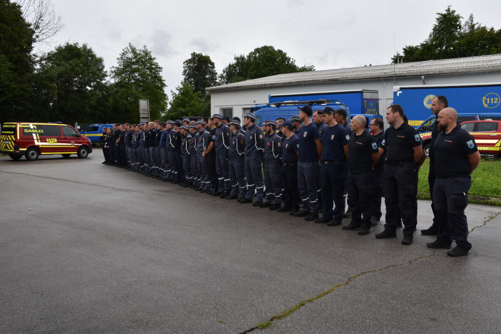 Pripadniki v modrih uniformah civilne zaščite in gasilski uniformi stojijo v postroju v dveh vrstah, slika iz desne.