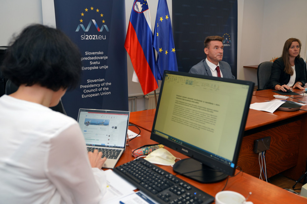 V ospredju oseba za računalnikom v ozadju še dve osebi za katerima sta slovenska in evropska zastava.