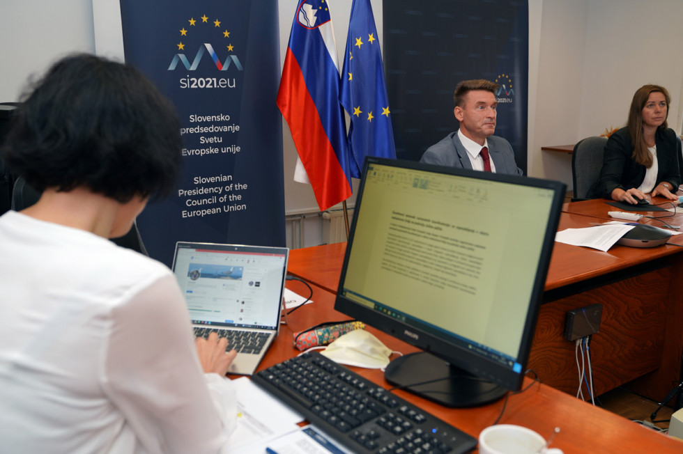 V ospredju oseba za računalnikom v ozadju še dve osebi za katerima sta slovenska in evropska zastava.