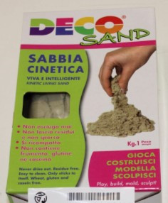 Na sliki je embalaža kinetičnega peska DECO SAND. Na njej je prikazana roka, ki sipa pesek, ter napisi o lastnostih izdelka. Pakiranje vsebuje 1 kg svetlo bež kinetičnega peska za igro, modeliranje in oblikovanje.