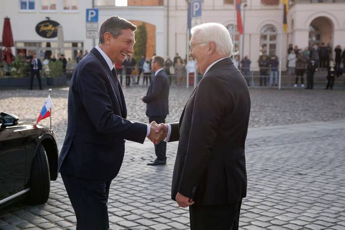 Die Präsidenten Pahor und Steinmeier schütteln sich auf dem Stadtplatz die Hände, im Hintergrund sind Menschen und Gebäude auf dem Hauptplatz zu sehen