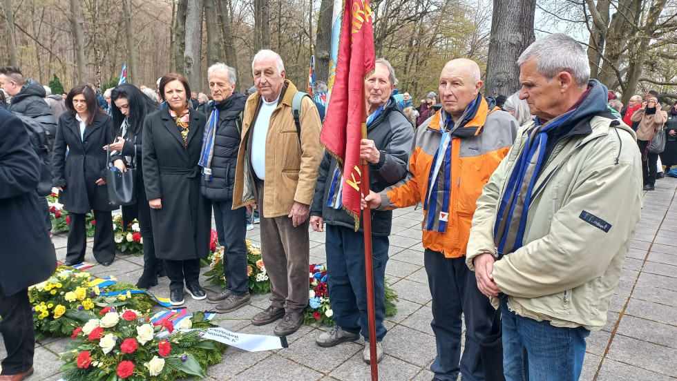 Spominska slovesnost na prostem s številnimi udeleženkami in udeleženci. Več starejših moških nosi modre rutke in drži rdečo zastavo. Pred njimi ležijo venci in cvetje v spomin na žrtve. Ljudje stojijo mirno in spoštljivo med visokimi drevesi v parku.
