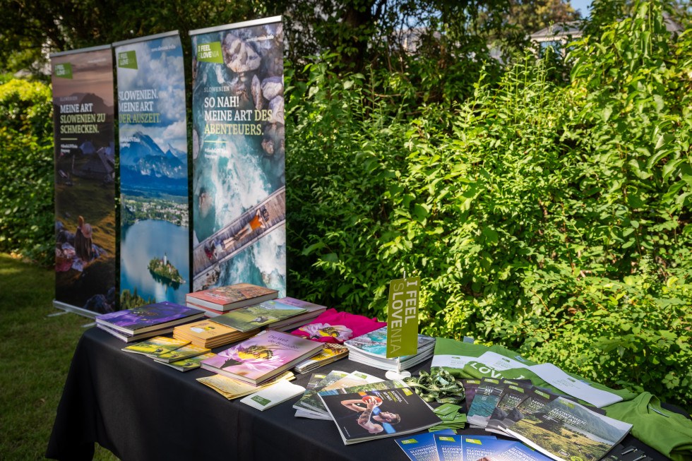 Ein Informationsstand über Slowenien im Garten. Auf einem Tisch liegen Broschüren, Bücher, Prospekte und Werbematerialien mit Informationen über Slowenien. Im Hintergrund stehen drei große „I feel Slovenia“-Banner mit Landschaftsmotiven. Der Stand ist von