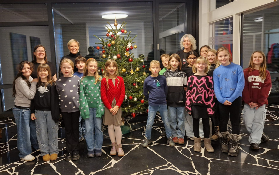 Ženska igra na kitaro, deklica na zvončke, ostali otroci pojejo zraven okrašene smreke.