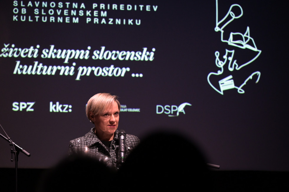 Ženska stoji in govori v mikrofon. Zadaj na temni podlagi napis: Slavnostna prireditev ob slovenskem kulturnem prazniku, živeti skupni slovenski kulturni prostor.