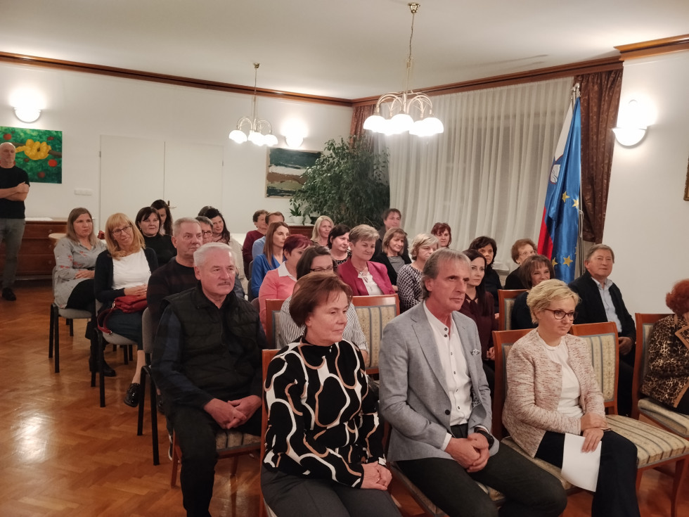 Udeleženci sedijo na stolih in poslušajo.