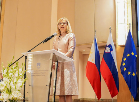 oseba govori za govornico, v ozadju zastave