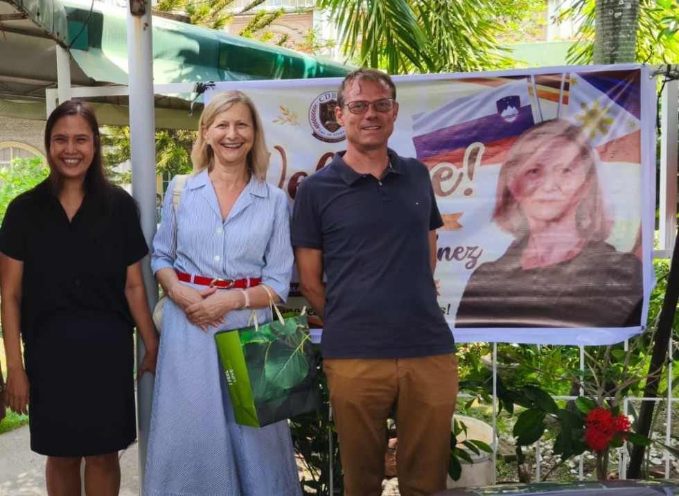 Three adults pose outdoors in front of a banner, two women and a man, all smiling.