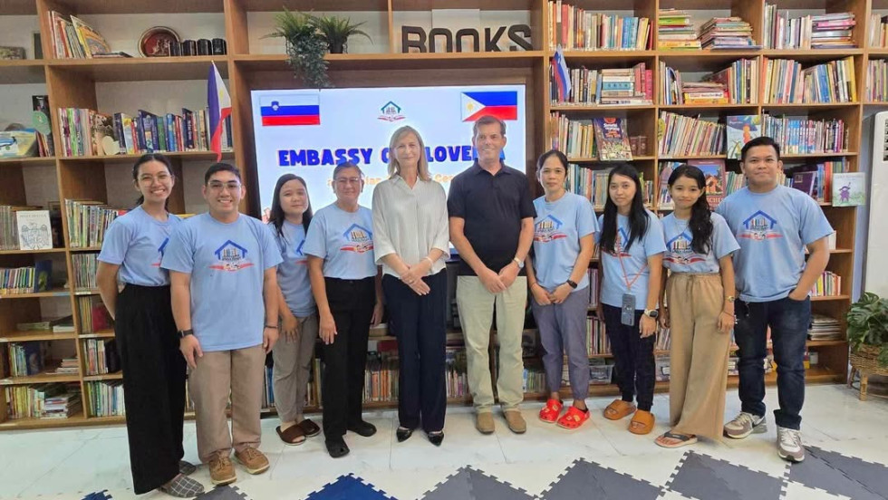 Group of ten in a library wearing matching light blue shirts, adults at center; screen behind reads 'Embassy' and flags.