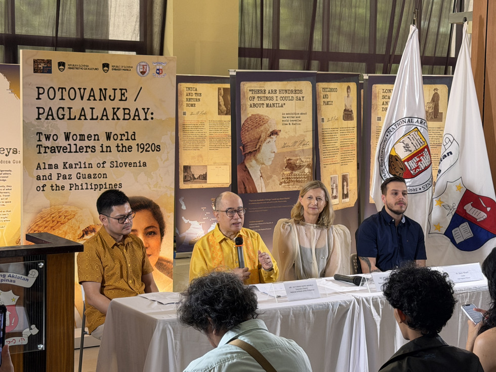 štiri osebe sedijo za mizo, ena drži mikrofon in govori. v ozadju razstavni paneli