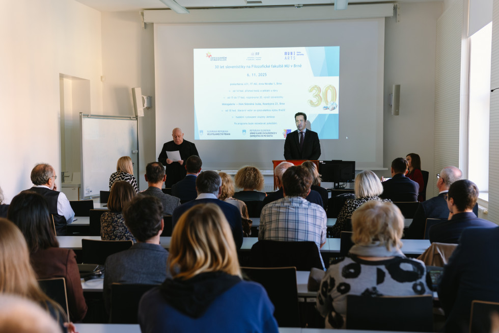 Učilnica z belimi stenami ima ob steni veliko projektno platno na katerem sveti projekcija. Pred platnom stojita dva gospoda srednjih let, oba elegantno oblečena v temne barve. Pred njima občinstvo, ki sedi v šolskih klopeh. Skupaj jih je približno 20.