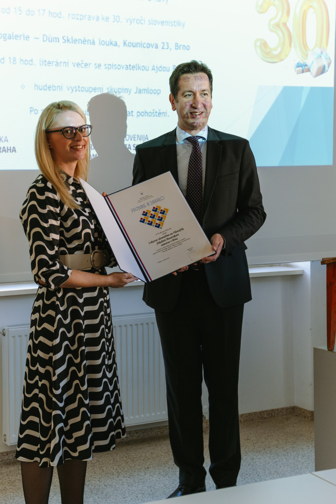 Na fotografiji sta dve osebi, ki stojita v svetlem prostoru pred projekcijskim platnom.  Levo stoji ženska svetlih las, oblečena v črno-belo obleko z valovitimi vzorci. Nosí očala. Desno stoji moški v temni obleki s kravato. Oba se nasmihata.