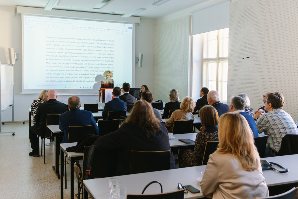 Na fotografiji je predavalnica. V ospredju sedijo udeleženci, večinoma odrasli. Obrnjeni so proti govornici. Govornica stoji za govorniškim pultom. Na projekcijskem platnu za njo je projicirano besedilo. 