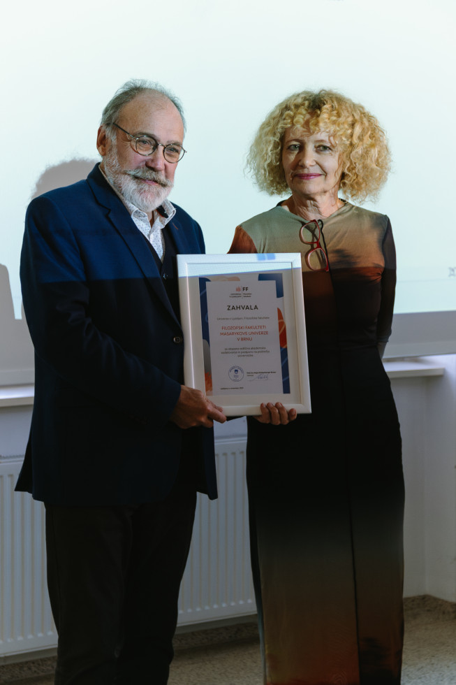 Na fotografiji stojita moški in ženska, oba sta obrnjena proti fotografu in se nasmihata. Moški drži okvirjeno listino. 