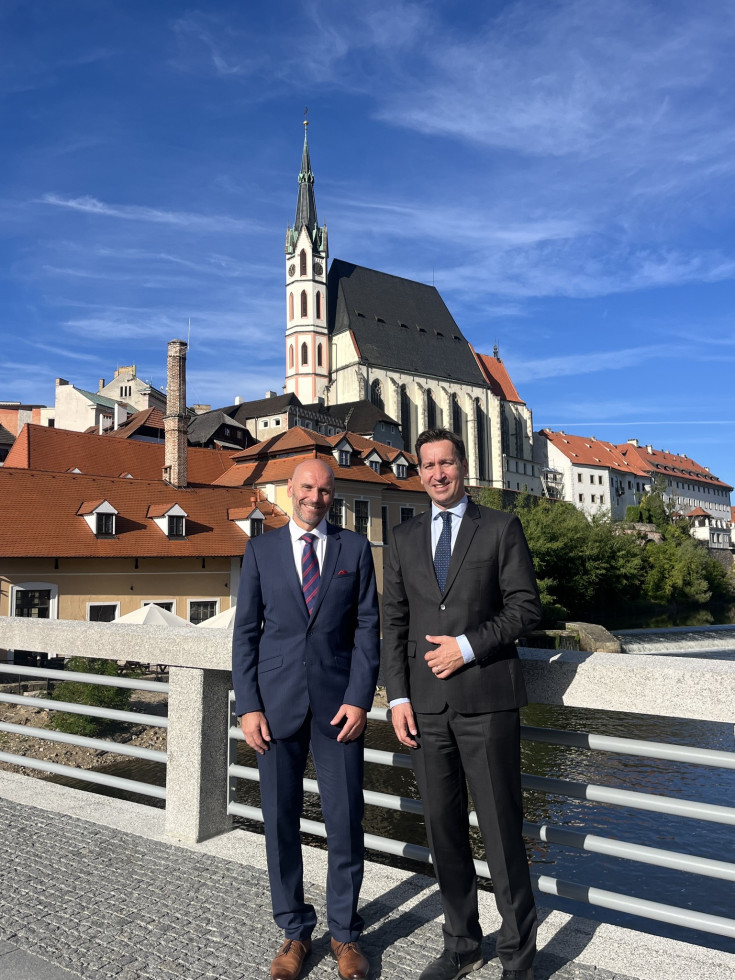 Dva moška, elegantno oblečena v suknjič s kravato stojita v sredini fotografije. V ozadju nad njima se dviga cerkev s ovalnim zvonikom, belo fasado in temno streho.