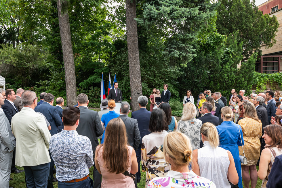 Množica ljudi v elegantnih oblačilih, obrnjenih s pogledom stran od fotografa, ki gledajo proti moškemu, ki stoji za govorniškim pultom v sredini fotografije. Moški je oblečen v črno obleko s skravato. Za njim so postavljene tri zastave na drogovih.