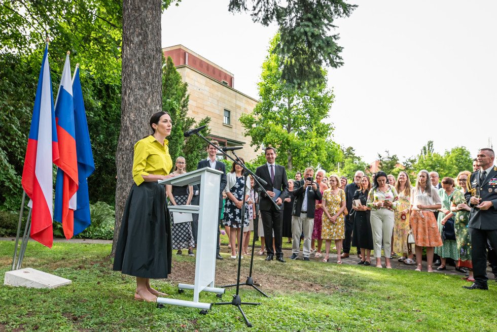 Na levi strani fotografija ženske za govorniškim pultom. Oblečena je v rumeno srajco in dolgo temno krilo. Za njo se nahaja drog s tremi zastavami v modrih, rdečih in belih barvah. Nekoliko v ozadju stojijo svečano oblečene oseba, tudi v vojaški uniformi.