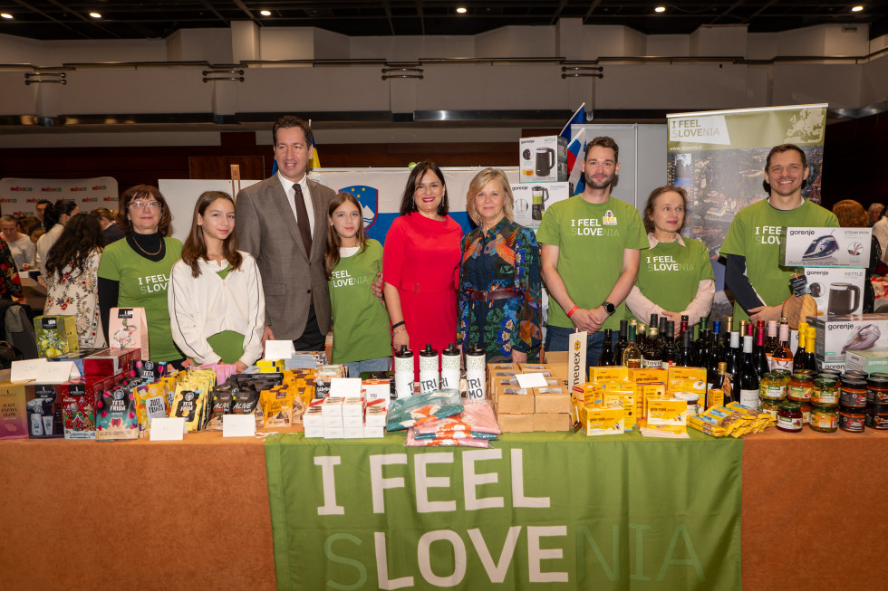 Na fotografiji skupina ljudi stoji za bogato obloženo stojnico z izdelki iz Slovenije, pred katero visi zelen prt z napisom »I Feel Slovenia«. Med izdelki so vidni čaji, čokoladne tablice, vino ter drugi slovenski izdelki, razporejeni v urejenih vrstah. V