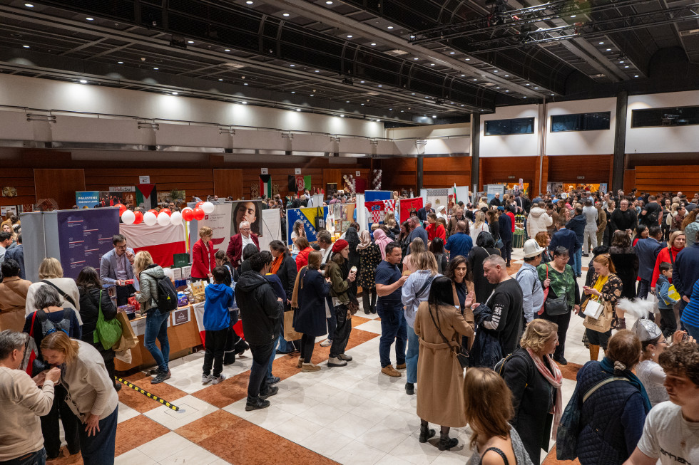 Na fotografiji je velika, živahna dvorana, polna obiskovalcev, ki se sprehajajo med stojnicami različnih držav. Ljudje klepetajo, nakupujejo in pregledujejo izdelke, vzdušje pa je praznično in družabno. Prostor je svetel in prostoren, z raznolikimi barvam