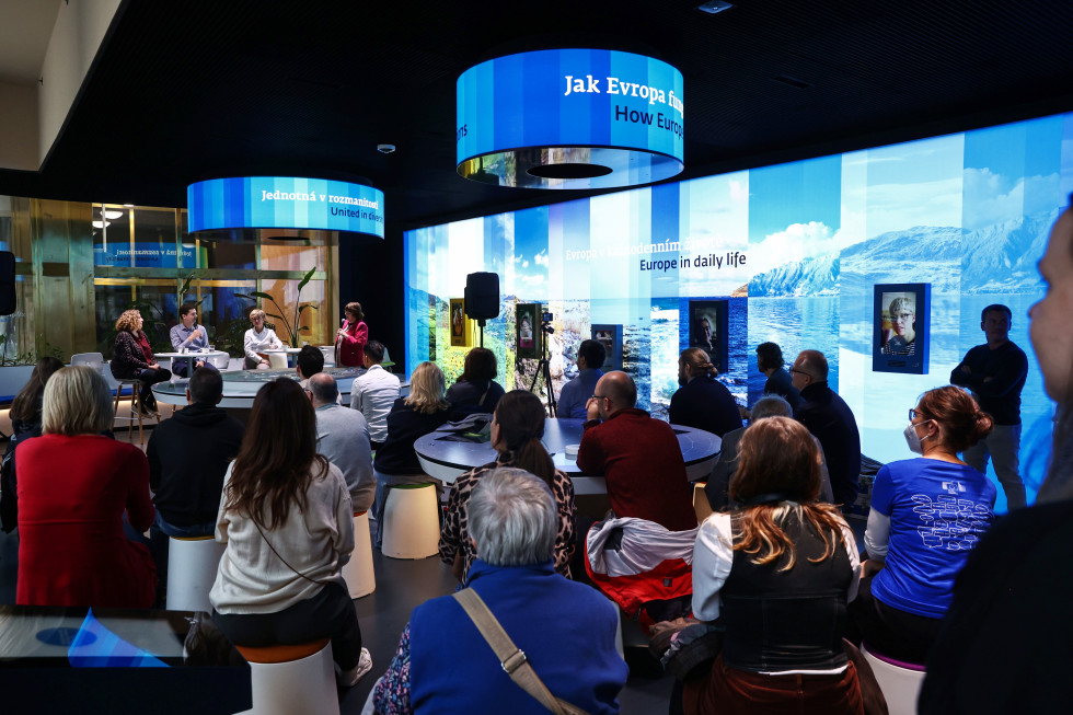 V ozadju fotografije pred občinstvom sedijo tri ženske in en moški. Ženska na desni strani mize in moški imata v roki mikrofon. Na desni strani sobe je čez celotno steno velik razsvetljen ekran v pretežno modri barvi z ozadjem pokrajine in portreti.  