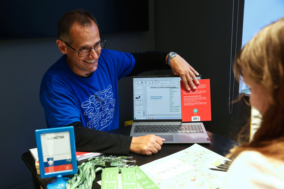 Za mizo sedi moški srednjih let z očali oblečen v modro majico. Z rdečo knjigo pokriva polovico ekrana računalnika. Na mizi so: knjižna kazala, vrvice za ključe, zemljevid Slovenije in stojalo z napisom "Slovinština",  pod katerim je slovenska zastava.