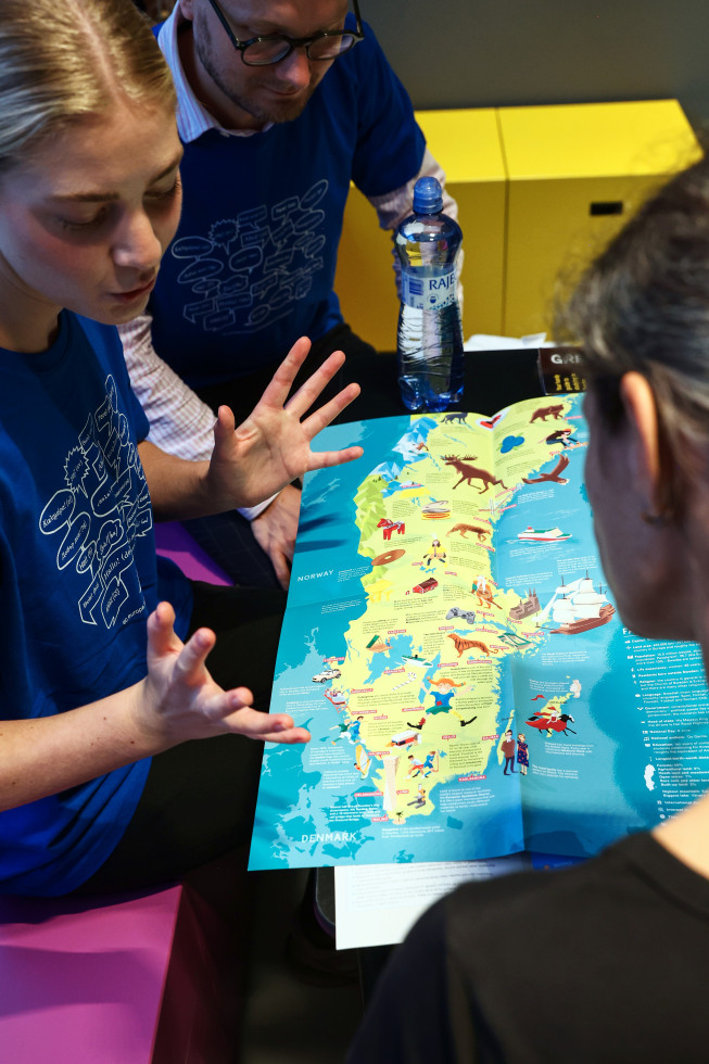 Na levi strani fotografije dekle z razprtimi rokami, pripoveduje obiskovalki, ki je na levi strani fotografije in vidimo le hrbtno stran. V zgornjem levem kotu sedi fant z očali. Na sredini fotografije zemljevid Švedske z različnimi slikami znamenitosti.